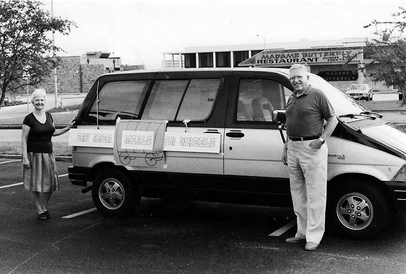 Two volunteers stand on either side of a van marked 'Bay Area Meals on Wheels' in a parking lot, early 1980s