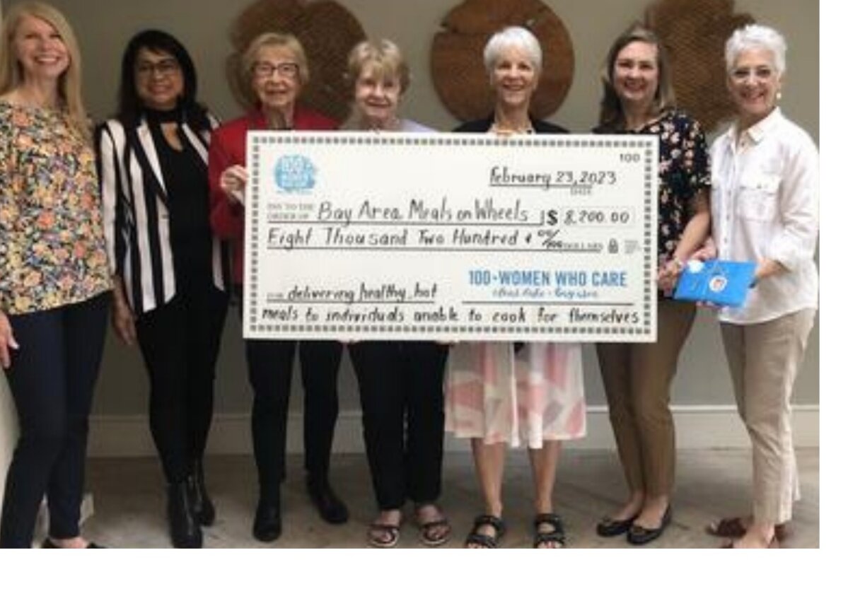 Seven members of 100+ Women Who Care presenting a large novelty check for $8,200 made out to Bay Area Meals on Wheels, dated February 23, 2023